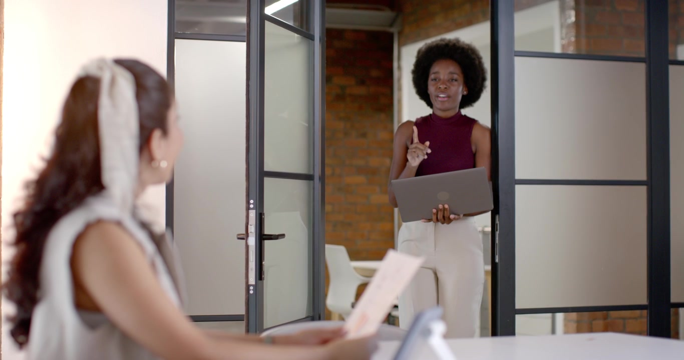 Diverse women talking as seated woman reading paper with tablet as peer entering pointing laptop