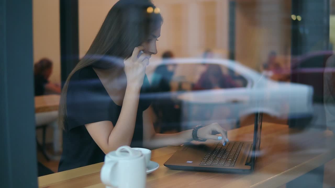 vista desde la calle de una joven atractiva hablando por teléfono en un café y mirando la pantalla de su computadora portátil durante