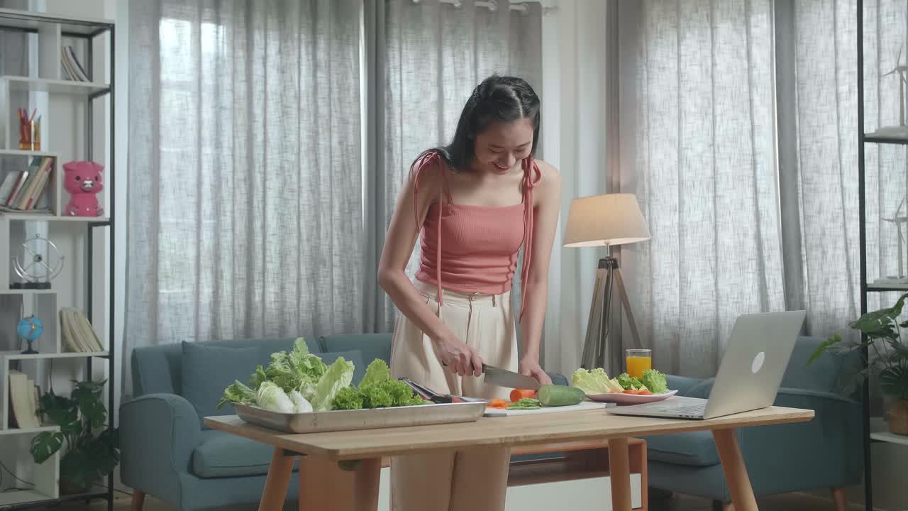 웃는 아시아 여성이 노트북으로 온라인에서 요리를 배우고 집에서 당근을 잘라내는 것을 즐긴다.