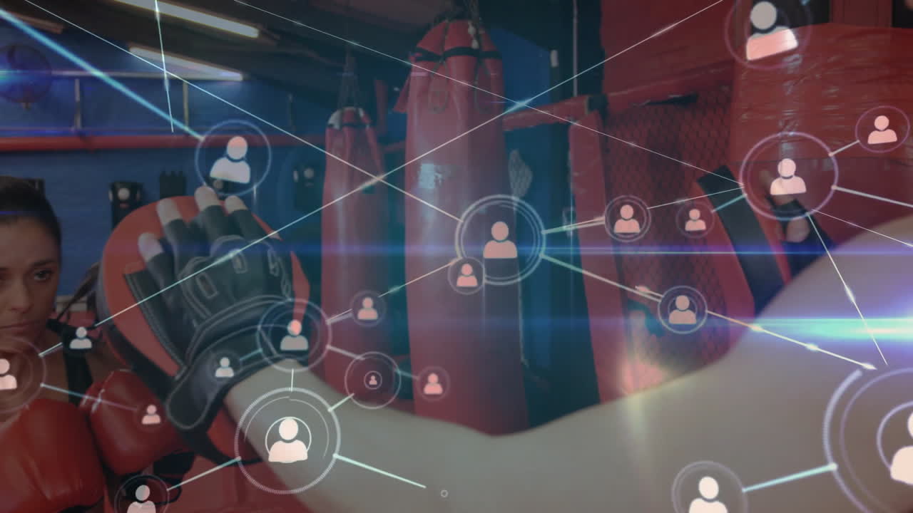 Teenage boy trainee boxer punching heavy bag in fitness gym, showing network icons connecting coach