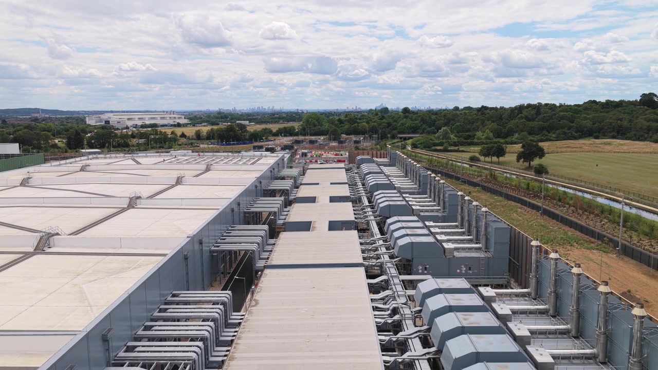 Rækker af industrielle køleenheder på taget af et datacenterkompleks på landet med grønne marker synlige under en overskyet himmel i de lyse dagtimer.