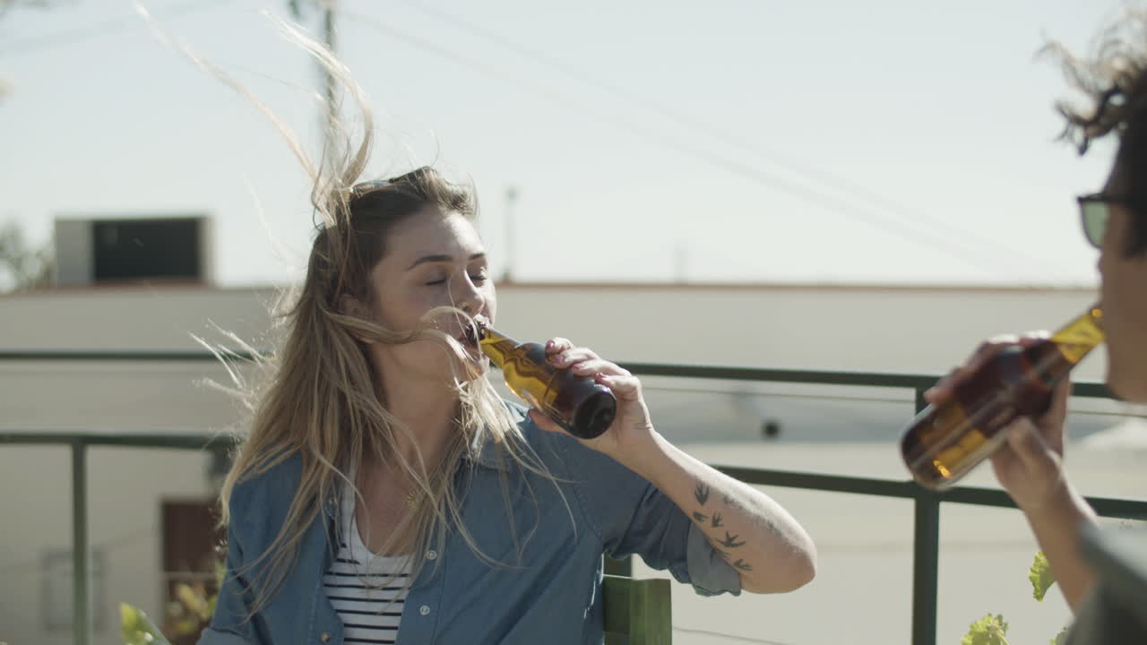 mujer feliz sentada en la azotea y tintineando botellas de cerveza con un amigo en un día ventoso