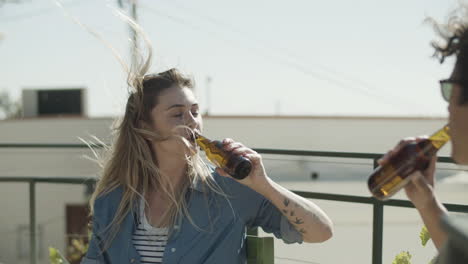 mujer feliz sentada en la azotea y tintineando botellas de cerveza con un amigo en un día ventoso