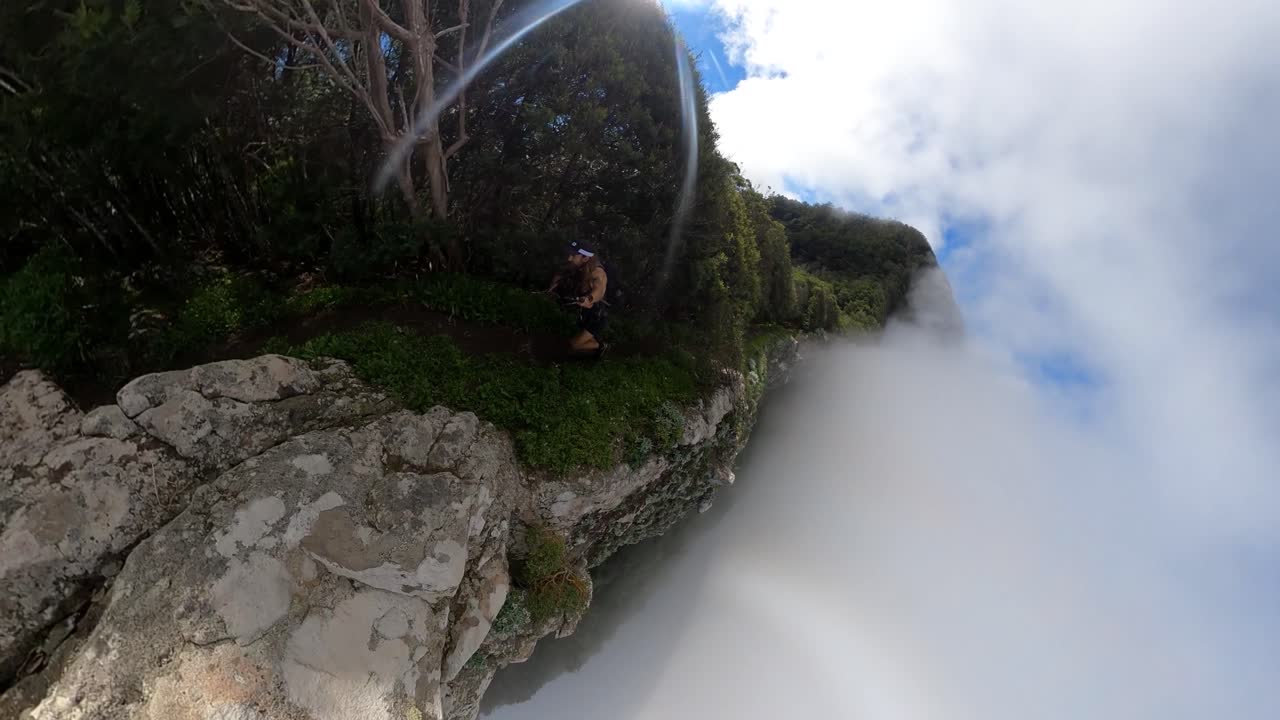 A young and strong solo hiker is walking along the steep edge of Espigao Amorelo in Madeira with thin clouds around him as he takes a dangerous step up to a rock on the cliffside.