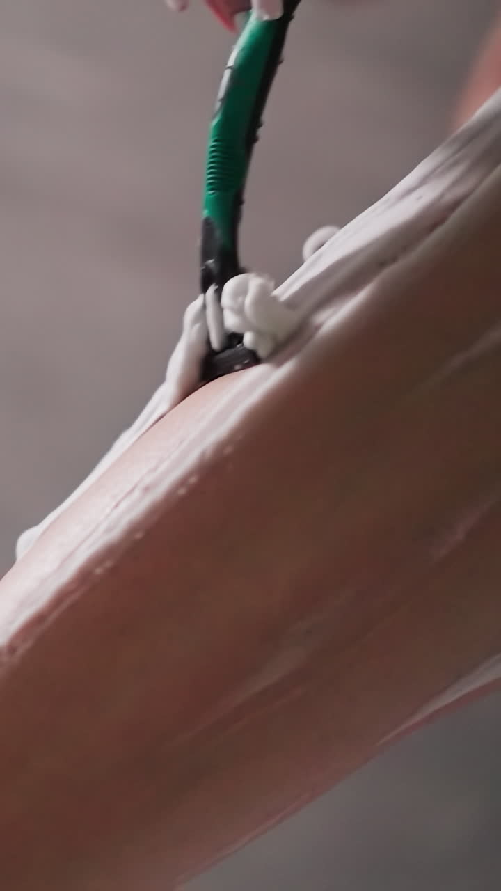 Woman with razor shaves leg hair in shower closeup. Lady removes body hair from skin covered with cream in bathroom. Woman does beauty routine