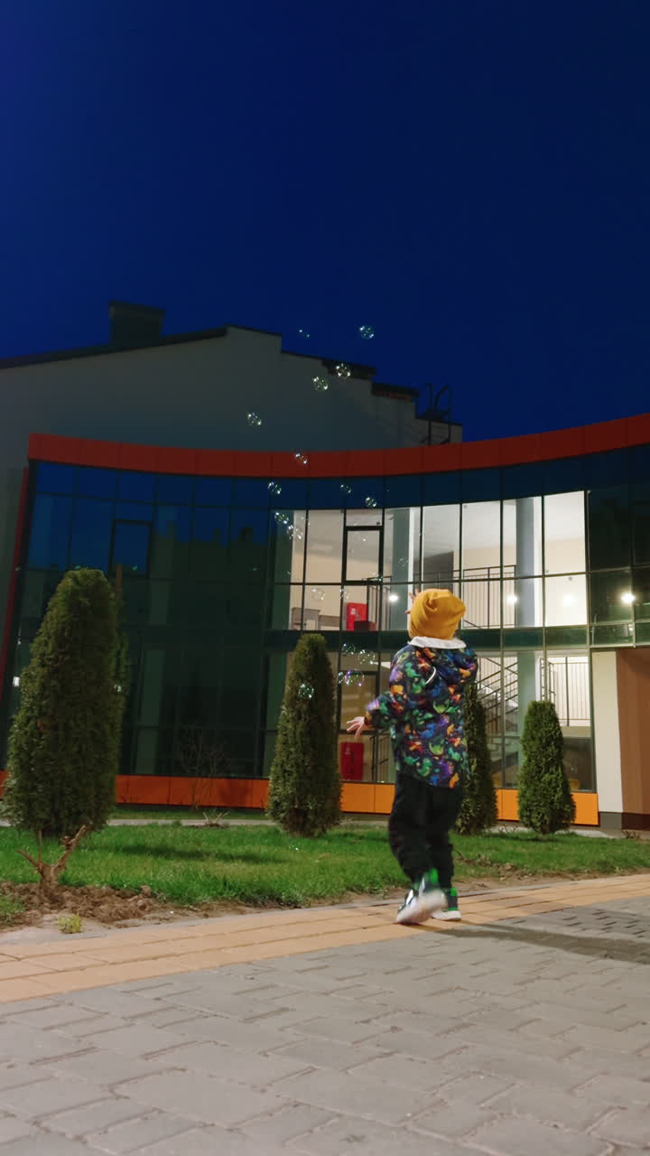 Male kid wearing jacket, cap and pants stands outdoors in the evening. Kid jumps happily and goes catching the soap bubbles. Low angle view. Vertical video.