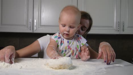 Young-mother-and-her-little-daughter-preparing-dough-on-the-table.-Little-baby-playing-with-flour.-Baker-prepares-the-dough