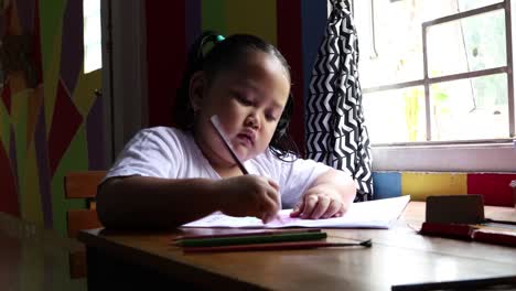 Cute-asian-little-girl-sitting-near-window-while-coloring-her-book