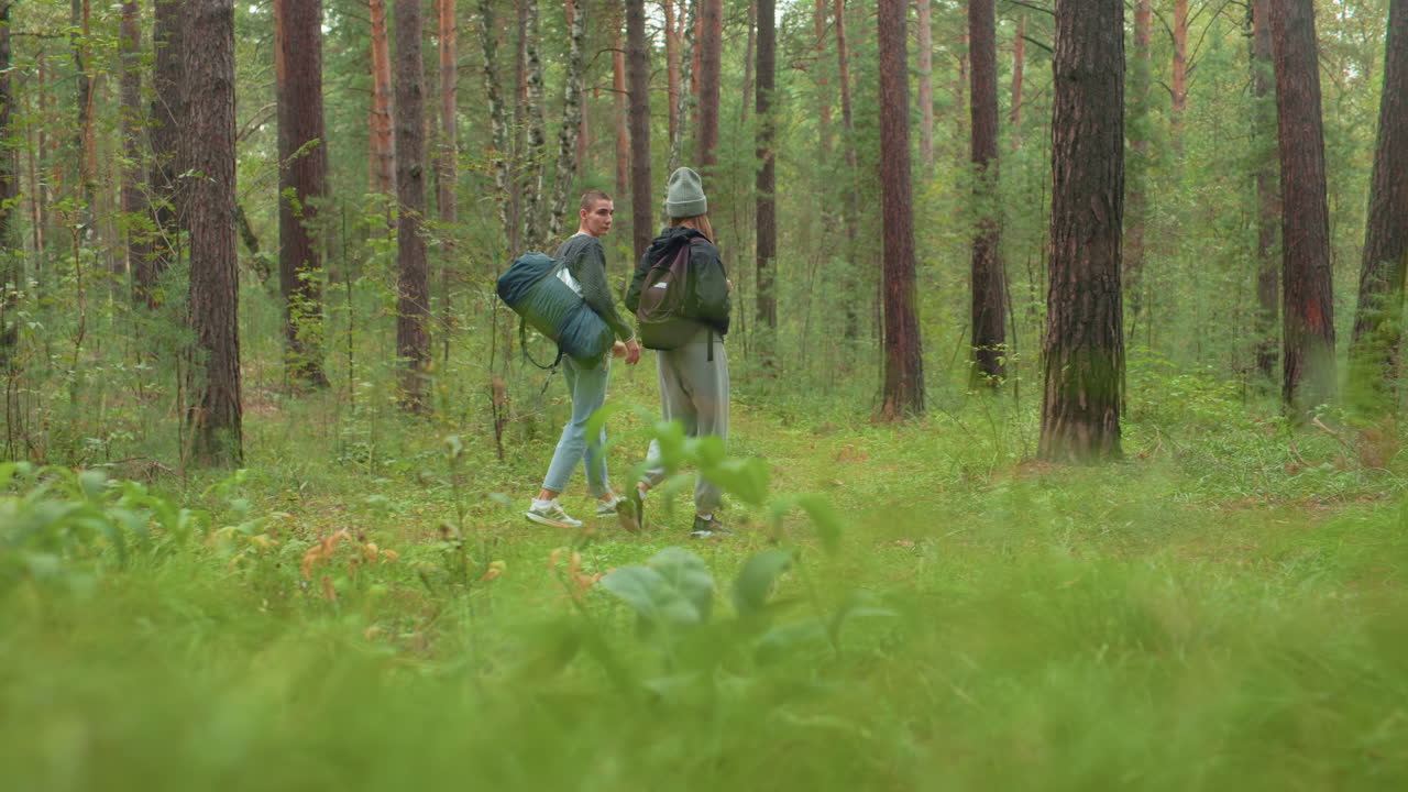 Couples standing in lush forest after dropping travel bags as man places hand on waist and looks around thoughtfully, surrounded by tall trees, dense greenery, and peaceful woodland atmosphere