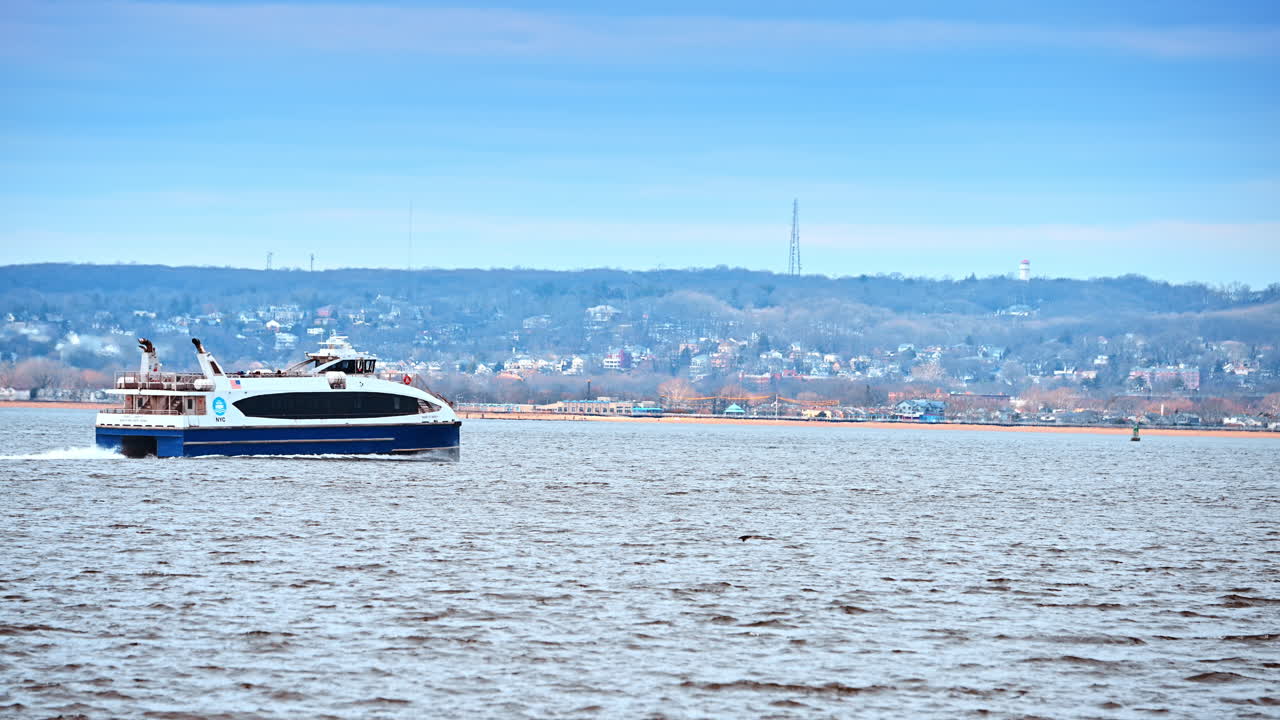 Traghetto passeggeri che naviga lungo la costa di New York. Un catamarano si muove attraverso le acque del porto con colline costiere e un faro visibili in lontananza.