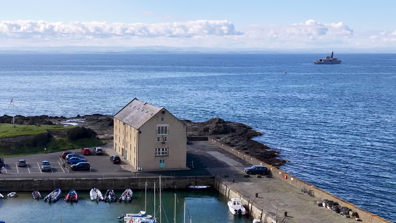 Drone glides over marina, boats, and historic pier building under bright daylight, revealing scenic coastline