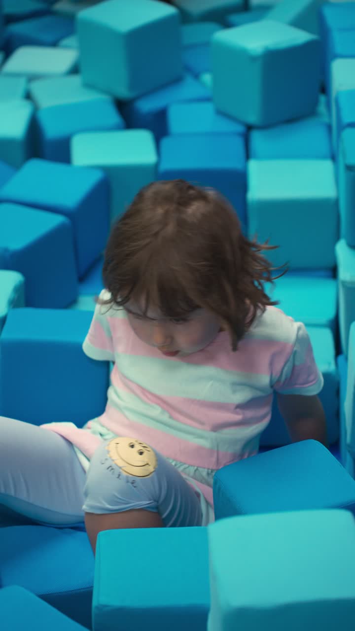 Adorable five year old girl lies on her back in a large blue foam cube pit and joyfully throws a soft block into the air in slow motion at an indoor playground