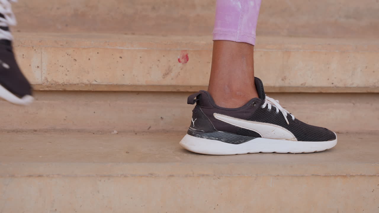 Primer plano de los pies de una atleta negra subiendo escaleras, mostrando una zapatilla negra sobre escalones de hormigón y leggings rosas visibles. El paso rítmico y el detalle de la suela, cordones y banda de rodadura transmiten movimiento y una vibra de entrenamiento urbano.