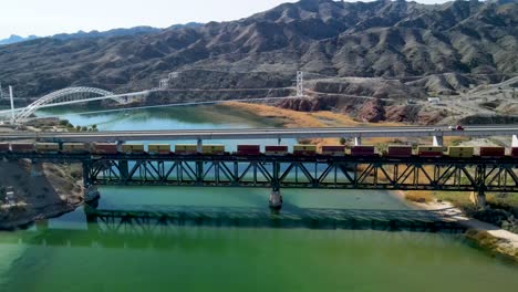 cargo train, crossing steel bridge over colorado river, the border between california and arizona, i-40 freeway east, wide angle establishing drone shot