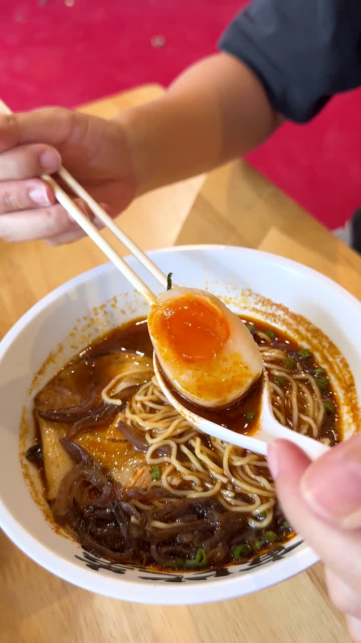 Una mano usa bacchette e un cucchiaio per sollevare un uovo alla coque dai ramen sotto una luminosa illuminazione da ristorante.