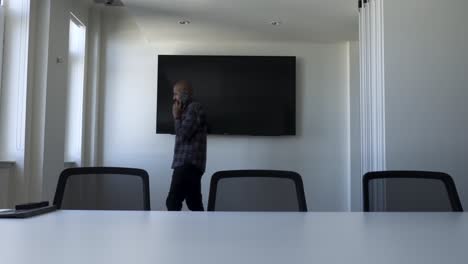 bald uk indian male talking on phone walking back and forth in modern empty office meeting room with large flat screen tv on wall