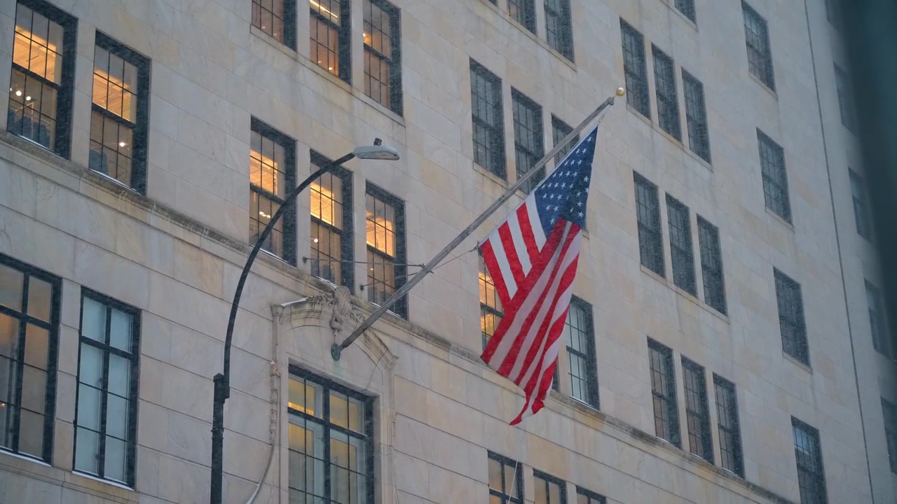 Bandiera americana su un classico edificio di Manhattan durante una tempesta di neve. Bandiera americana su asta montata sulla facciata di un edificio in pietra beige, nevicata, finestre illuminate, lampione, vista dal basso.