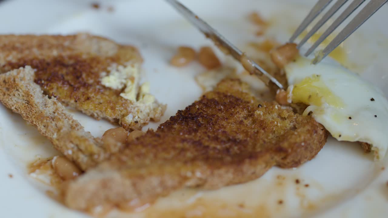 Fork and knife cutting toast, eggs, and beans on breakfast plate in natural lighting