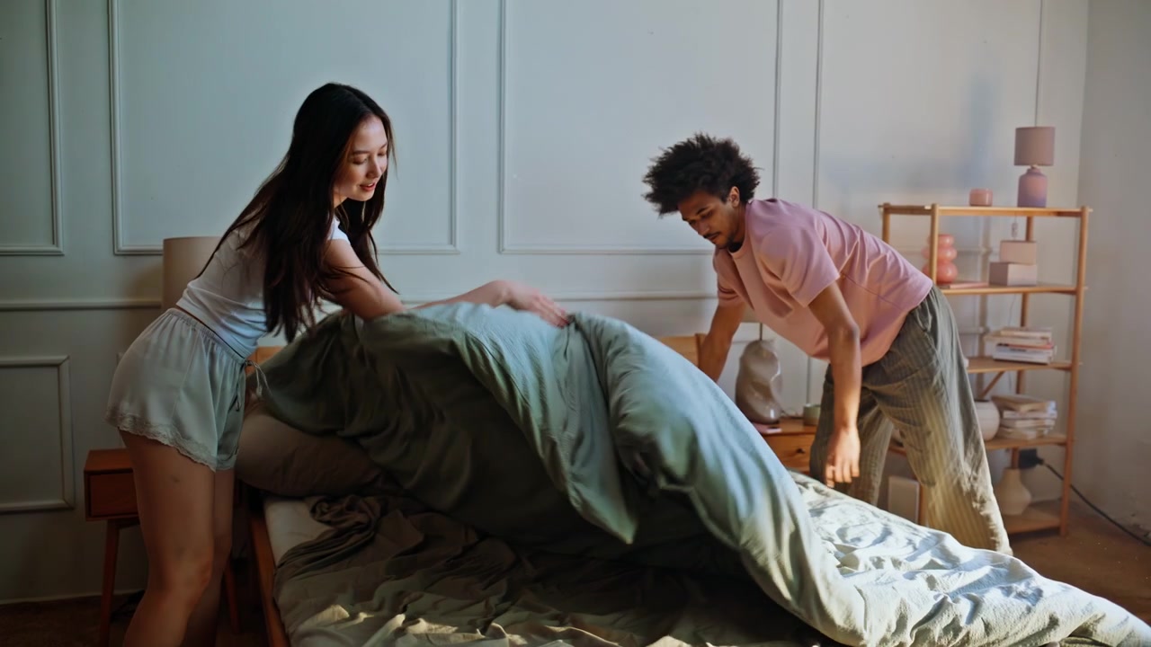 Partners making bed together morning. Interracial pair changing bedding home.