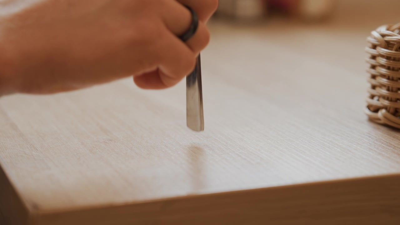 Close up of hand holding and moving metal tool over wooden surface. Hand with ring interacts steadily creating impression on wood while wicker basket remains in background. Soft lighting