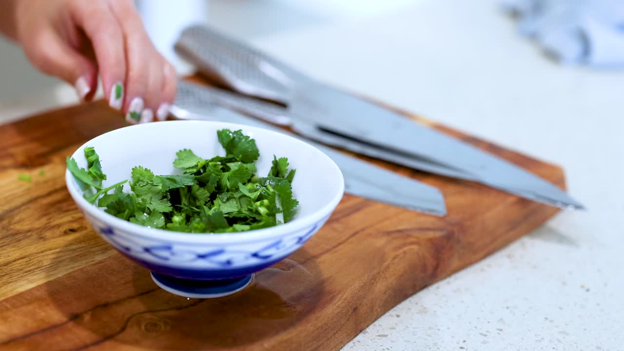 A person chops and arranges fresh coriander in a bowl on a wooden cutting board under bright kitchen lighting