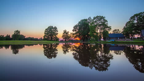 Tiro-De-Lapso-De-Tiempo-De-La-Puesta-De-Sol-En-El-Fondo-Y-La-Cabaña-Al-Lado-De-Un-Lago-En-Primer-Plano-Al-Atardecer