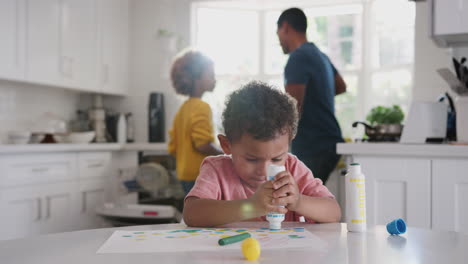 Junger-Afroamerikanischer-Junge-Malt-Ein-Bild-In-Der-Küche,-Während-Vater-Und-Schwester-Im-Hintergrund-Essen-Zubereiten