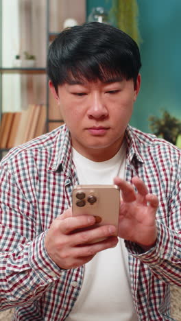 Asian man reading important message on smartphone pausing scroll tense thoughtful reaction on sofa