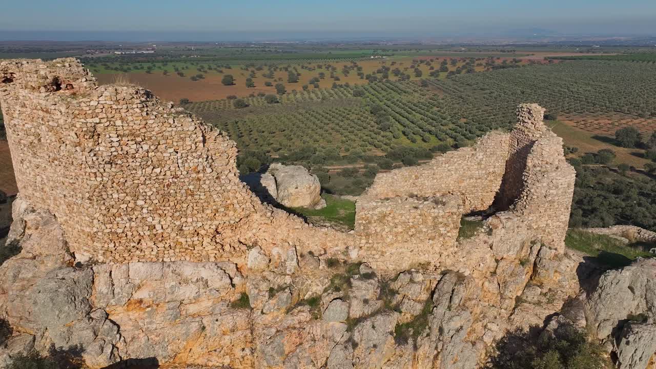 Drone reverse flight over the ruins of Dos Hermanas Castle in Navahermosa, Toledo. The castle stands on a rocky hill with olive groves, farmland, and mountains in the distance