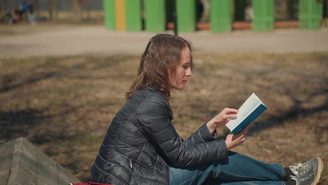 Close up of woman reading book outdoors on wooden bench with red bag nearby. Book is open and held in hands showing text details. Background includes green structure trees pathway soft