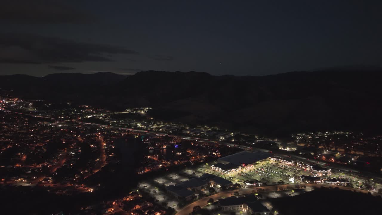 Sorvolo aereo notturno di Agoura Hills, California, USA, con traffico luminoso, centro commerciale illuminato e quartieri residenziali circostanti con luci della città che si estendono attraverso il paesaggio della valle