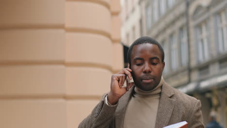 Vista-Cercana-Del-Hombre-Afroamericano-Mirando-Su-Cuaderno-Abierto-Y-Hablando-Por-Teléfono-En-La-Calle-En-Otoño