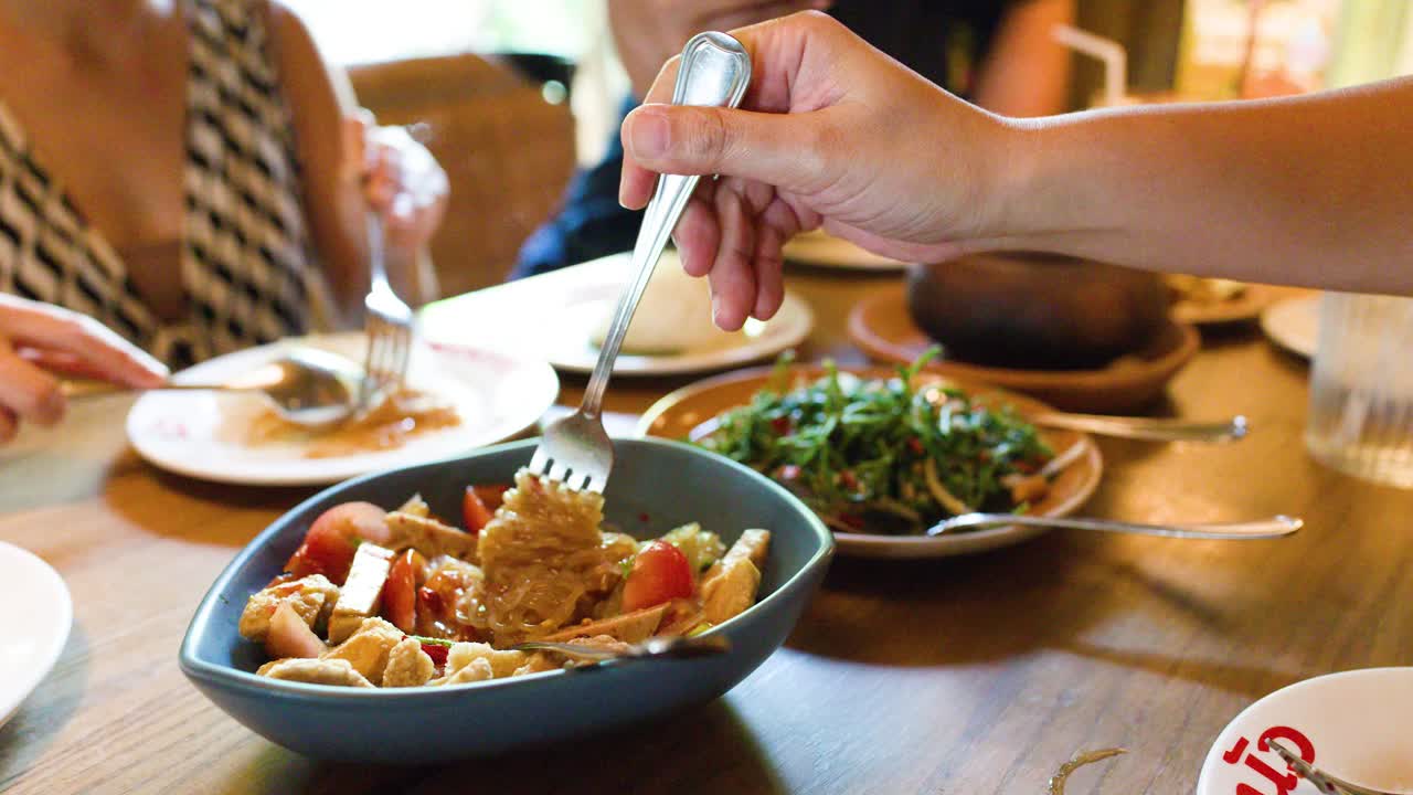 Hand lifts spicy rice vermicelli salad with fork, natural daylight, casual Bangkok restaurant dining scene