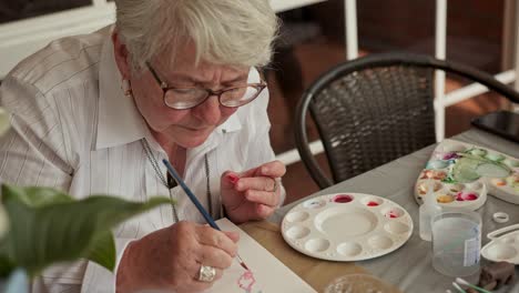 Older-grandma-looks-down-as-she-brushes-fine-strokes-of-blue-watercolor-paint-across-paper-pad