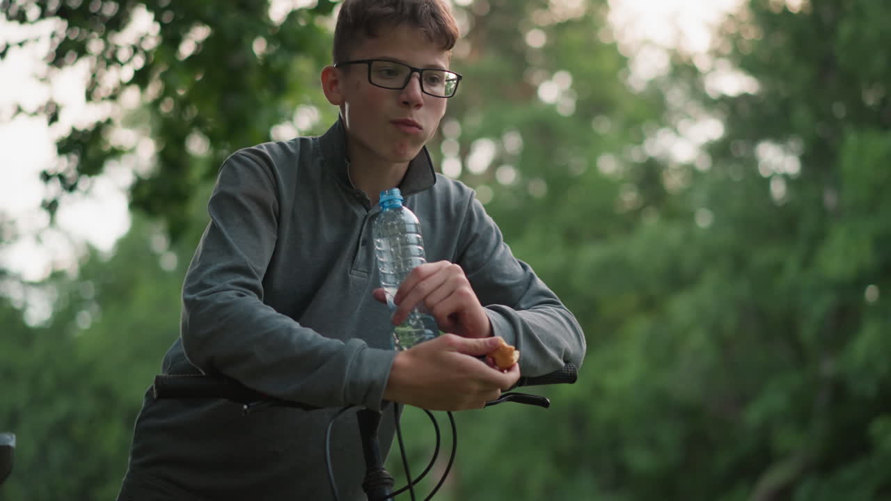 un niño en la parte superior gris se apoya contra el manillar de la bicicleta mientras saborea un bocadillo sabroso, sosteniendo una botella de agua en su otra mano, el fondo presenta un suave borrón de follaje verde