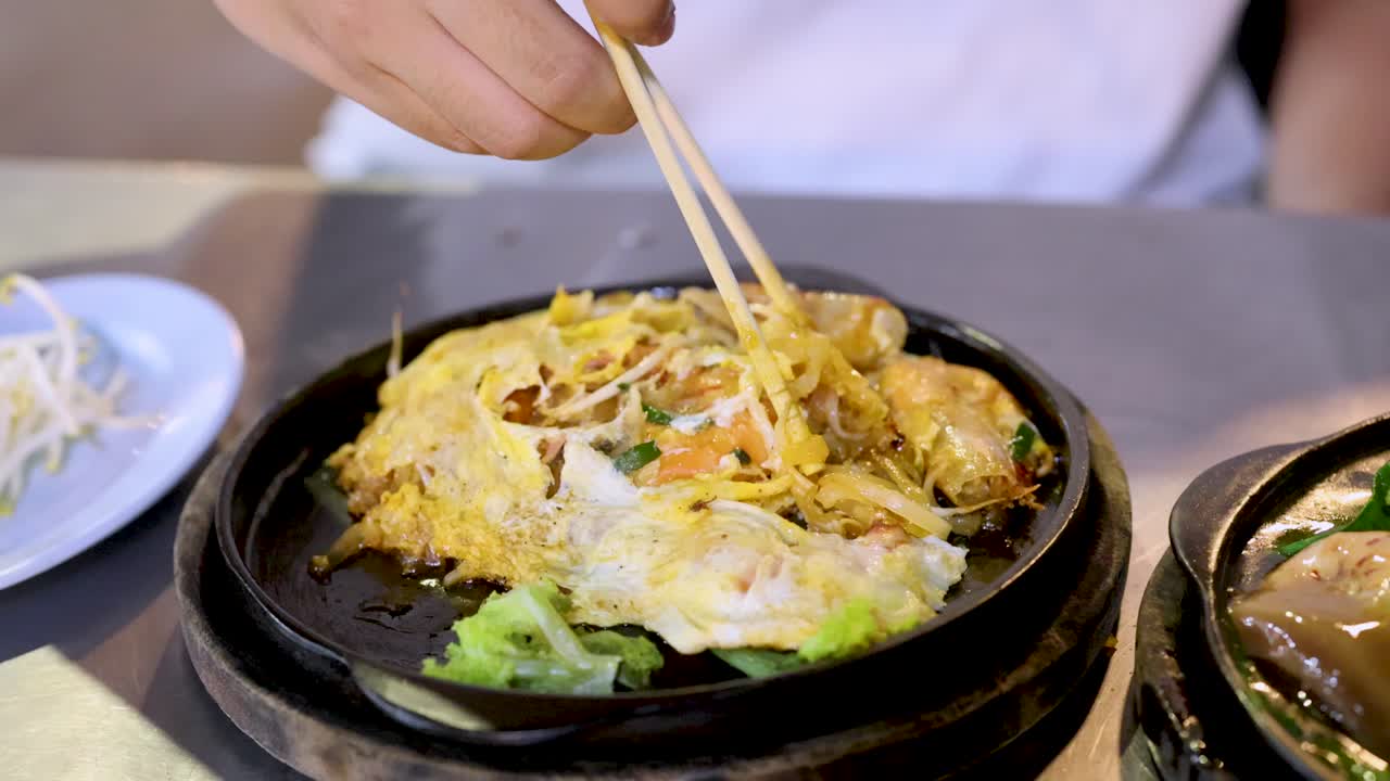 Person lifts hot plate Pad Thai noodles with chopsticks, bright lighting, close-up, casual dining