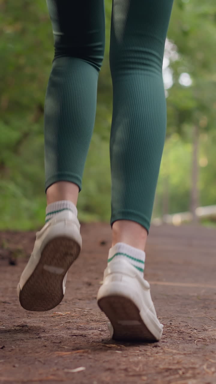 Female jogger running along path in park. Woman legs in sports shoes running on pathway surrounded by trees. Lady training in nature on summer day