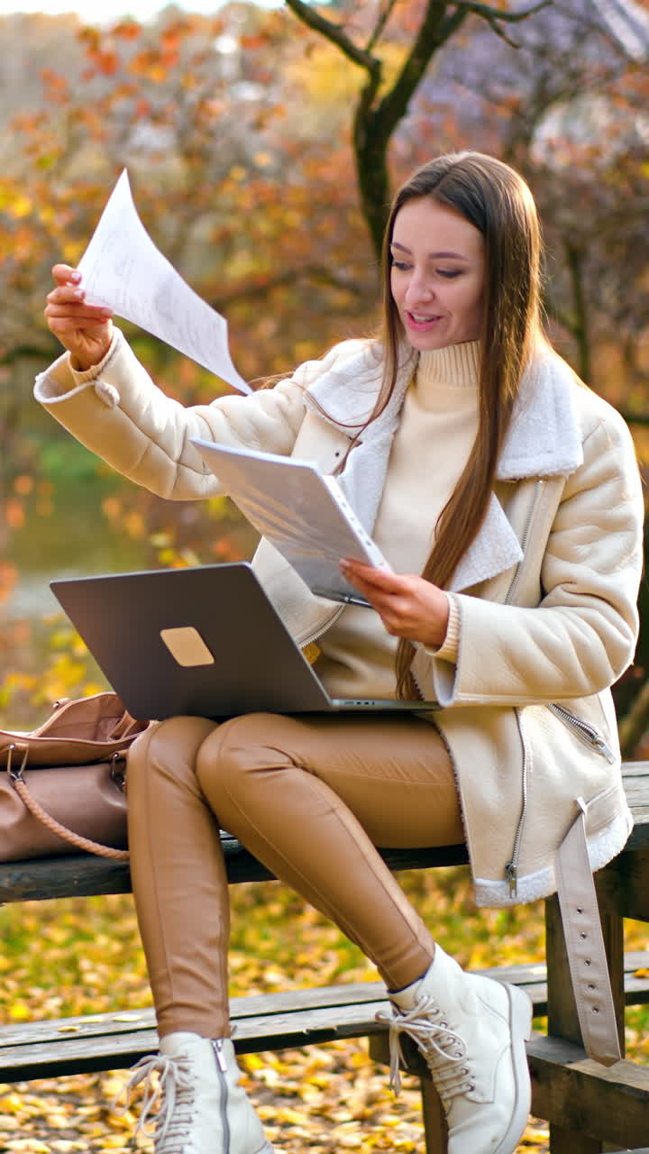 Happy woman working remotely in the autumn park. Lady checking up the documents and holding computer on her laps. Blurred backdrop. Vertical video
