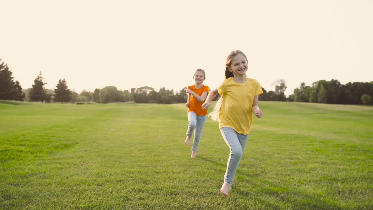 Two Little Sisters Running Barefoot Towards Camera On Meadow In The Park