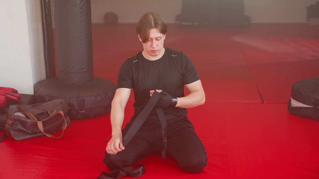 Athlete prepares for training session on red mat inside gym, wearing black sportswear and wrist wraps, showing focus, determination, and discipline while seated in readiness for boxing
