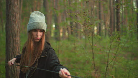 young lady wearing beanie focused on connecting flexible tent pole in forest, surrounded by tall trees and greenery, dressed in dark jacket, engaged in calm and intentional outdoor camping setup