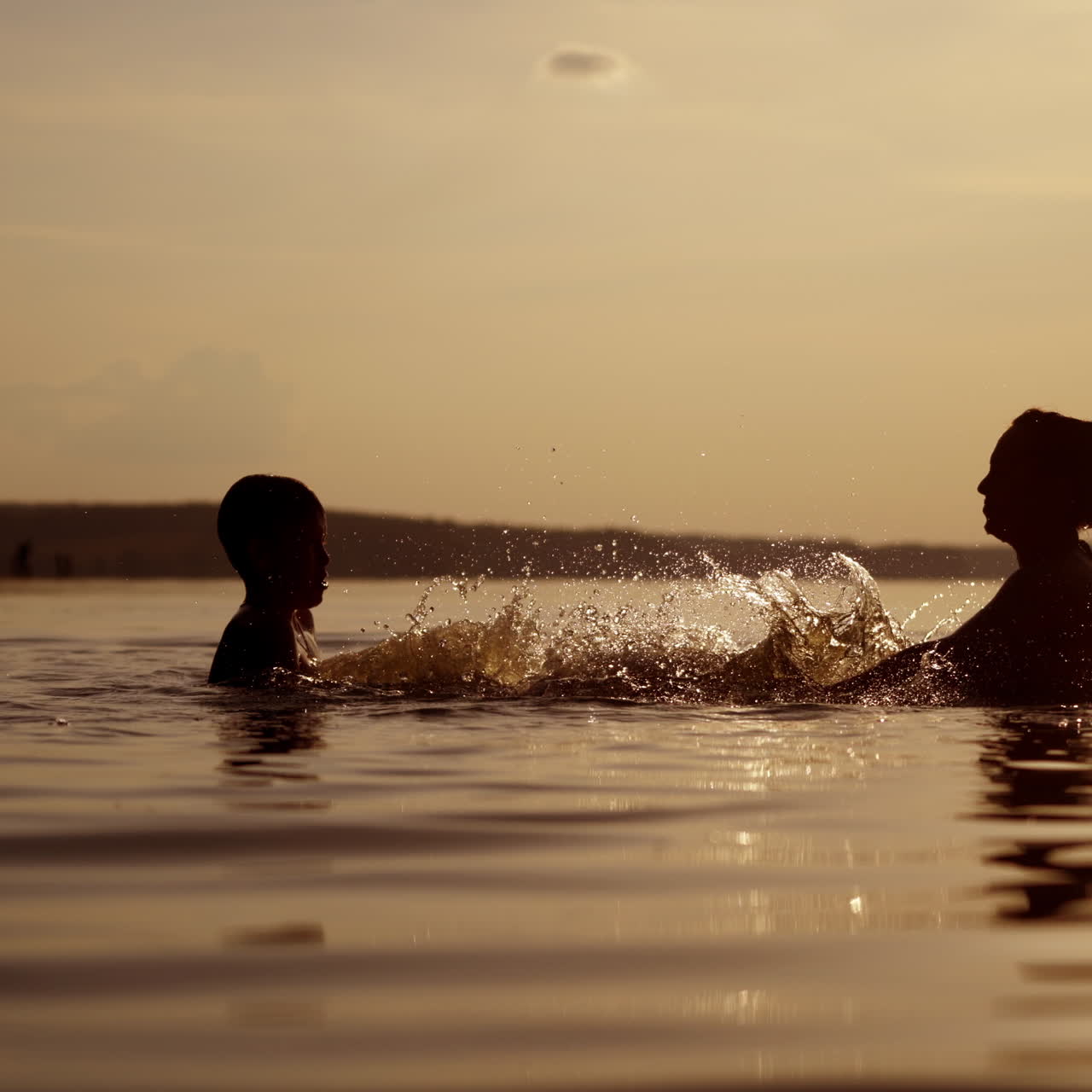 Silhouette of a woman and a boy inside the river at dusk. Mother and son making splashes on water happily in the evening.
