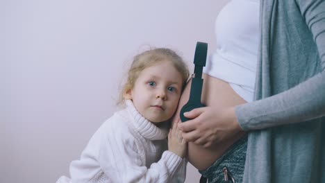 chica divertida pone la cabeza en el estómago de la mamá embarazada con auriculares