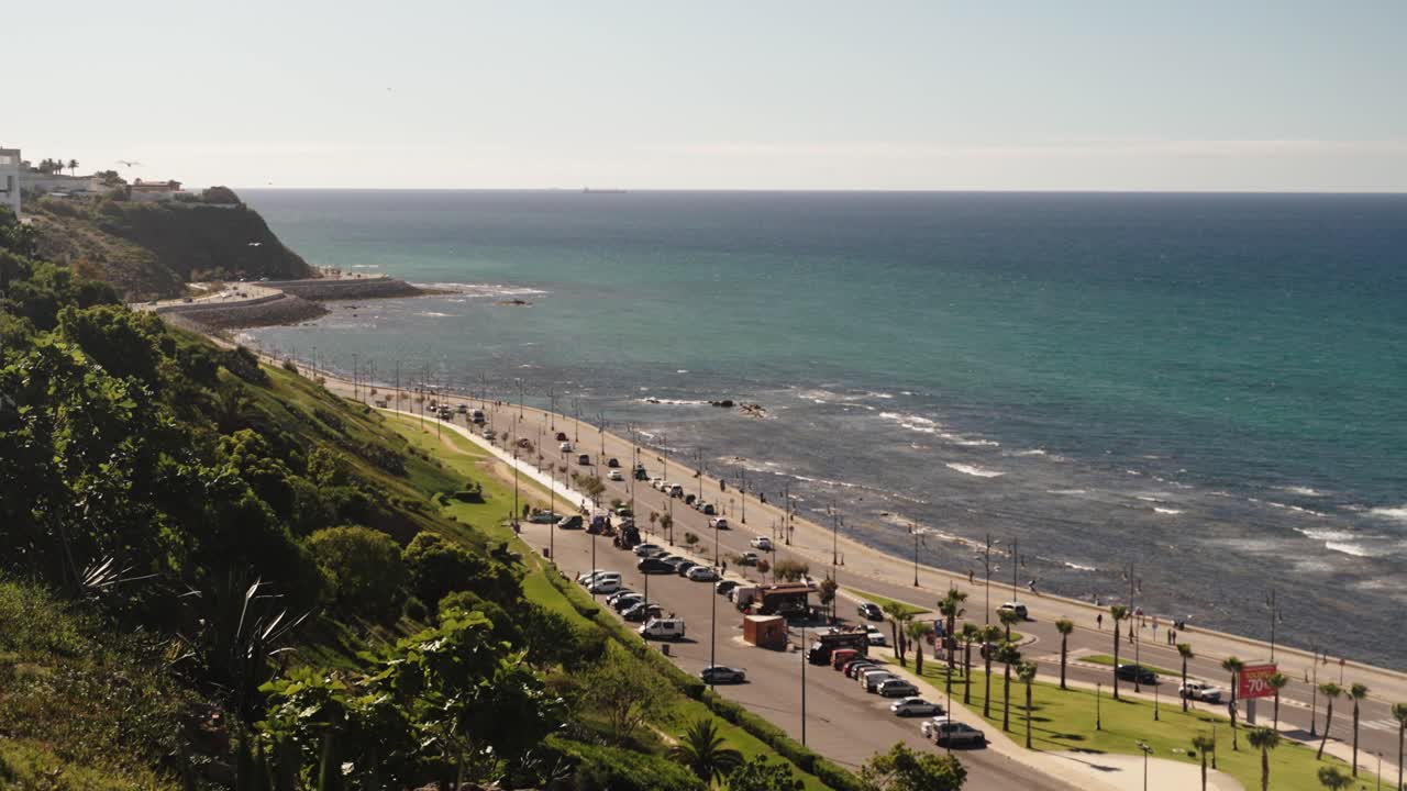 Cars drive along the scenic coastal road of Tangier, Morocco, with the turquoise sea stretching beside the city shoreline