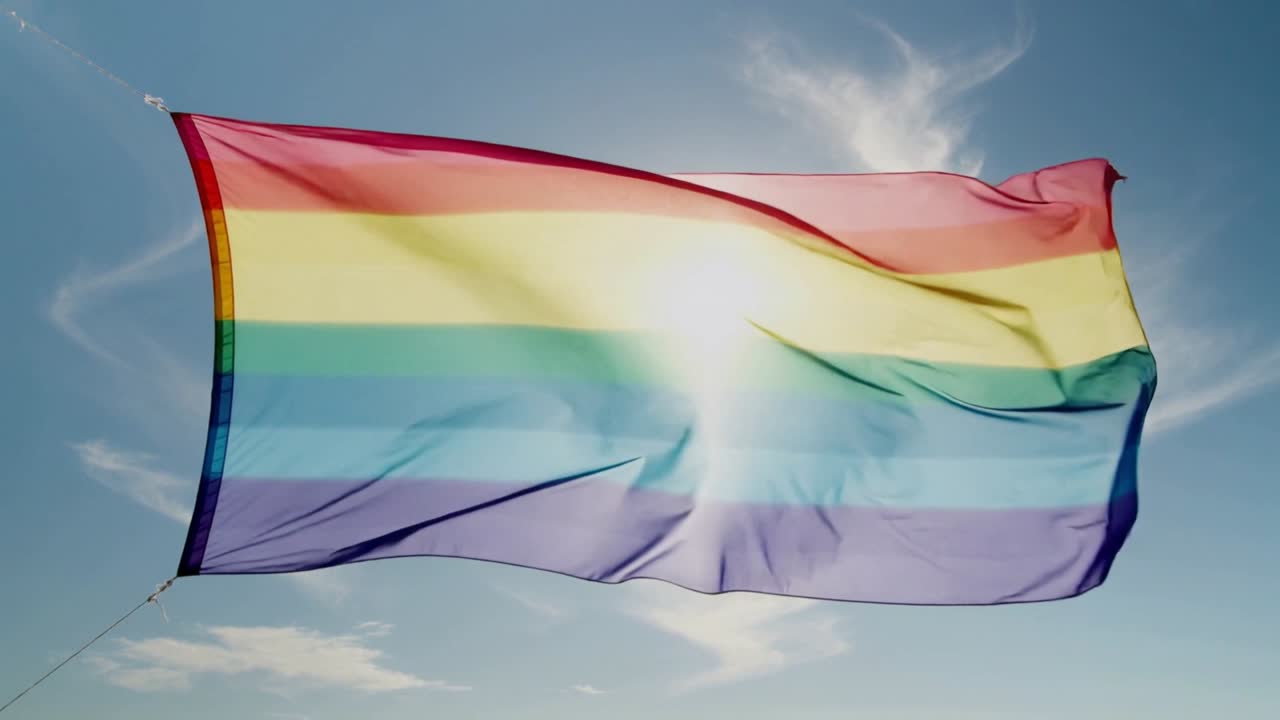 Grande bandiera arcobaleno ondeggiante che sventola nel cielo blu per la brezza, con il sole che retroillumina i punti di ancoraggio.