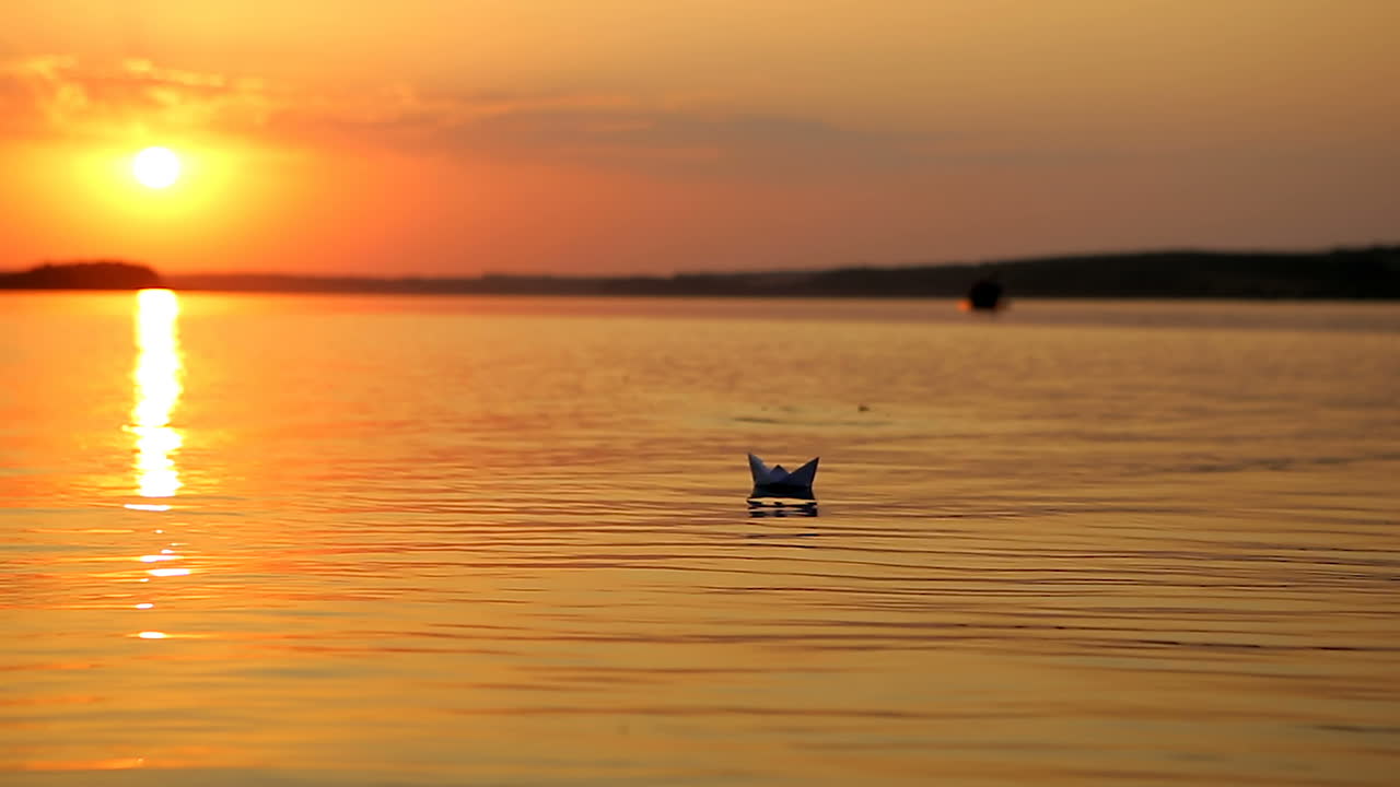 Paper Boat Sailing At Sunset. Origami paper boat sailing on blue water surface at sunset
