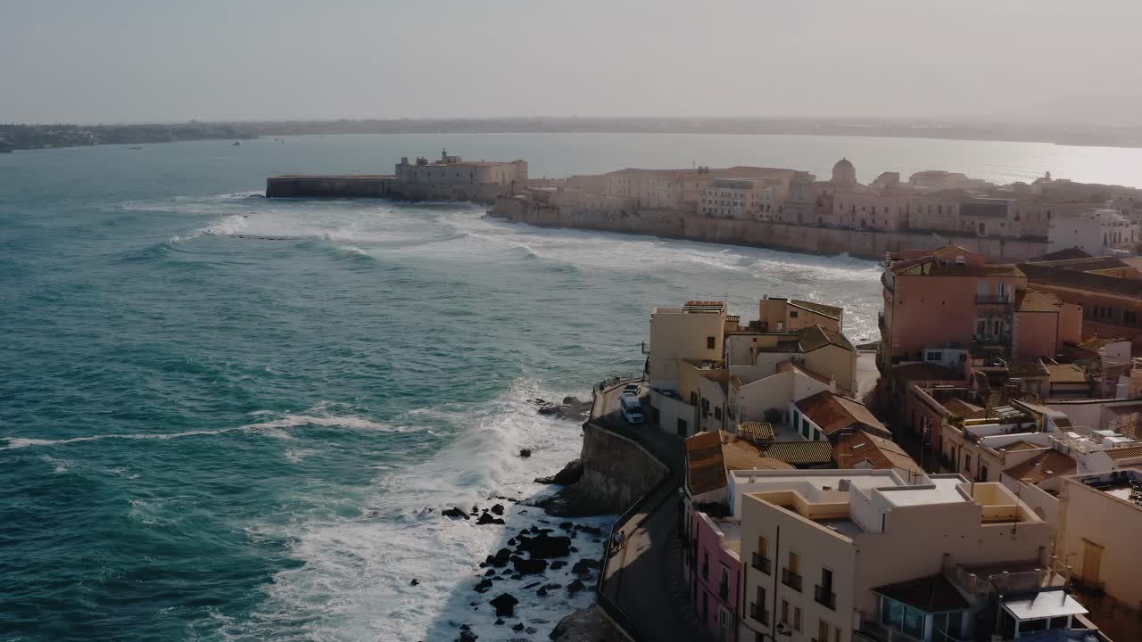 Aerial drone shot of Ortigia island, the old town of Syracuse in Sicily. Historic buildings by the Mediterranean sea with large waves crashing at the shore. Natural fortress around the island. UNESCO.