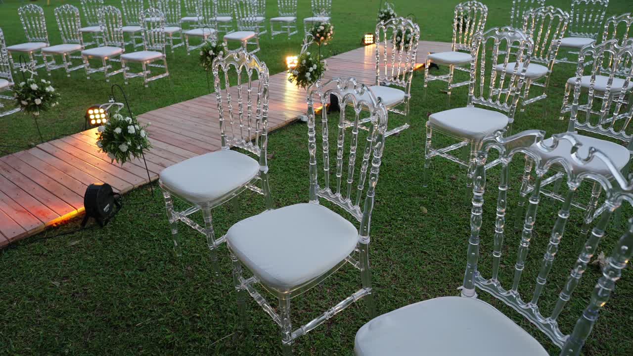 Allestimento per una cerimonia di matrimonio all'aperto con sedie trasparenti sull'erba, corridoio decorato con composizioni floreali e illuminazione soffusa, creando un'atmosfera per l'evento raffinata e romantica.