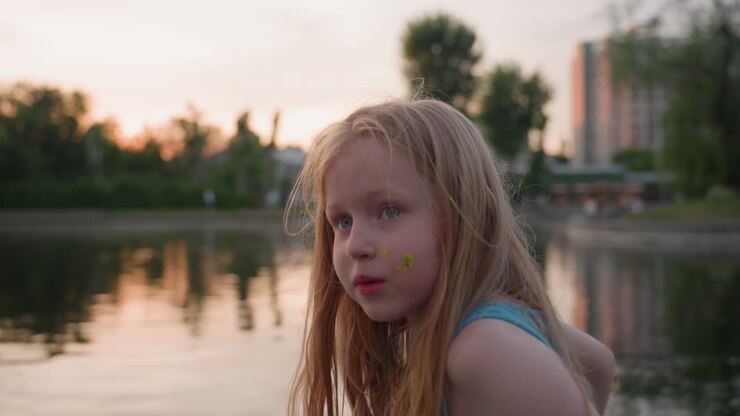 jeune enfant joyeux en extérieur au coucher du soleil sur un pont au bord de la rivière, taches de peinture sur la joue, expression espiègle tout en touchant ses cheveux, débardeur bleu décontracté, éclairage chaud de l'heure dorée et moment de joie spontané