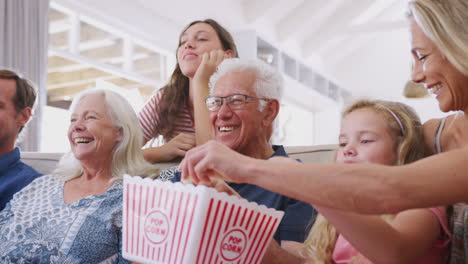 multi-generation family sitting on sofa at home eating popcorn and watching movie together